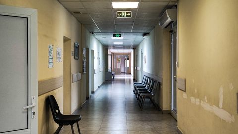 An empty hospital corridor with open doors and chairs, lit by fluorescent lights.