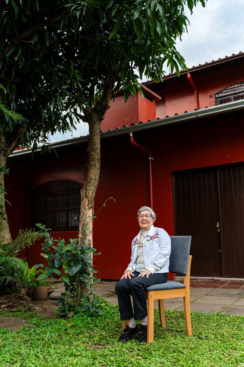Free Elderly woman seated outdoors in garden beside a red house. Stock Photo