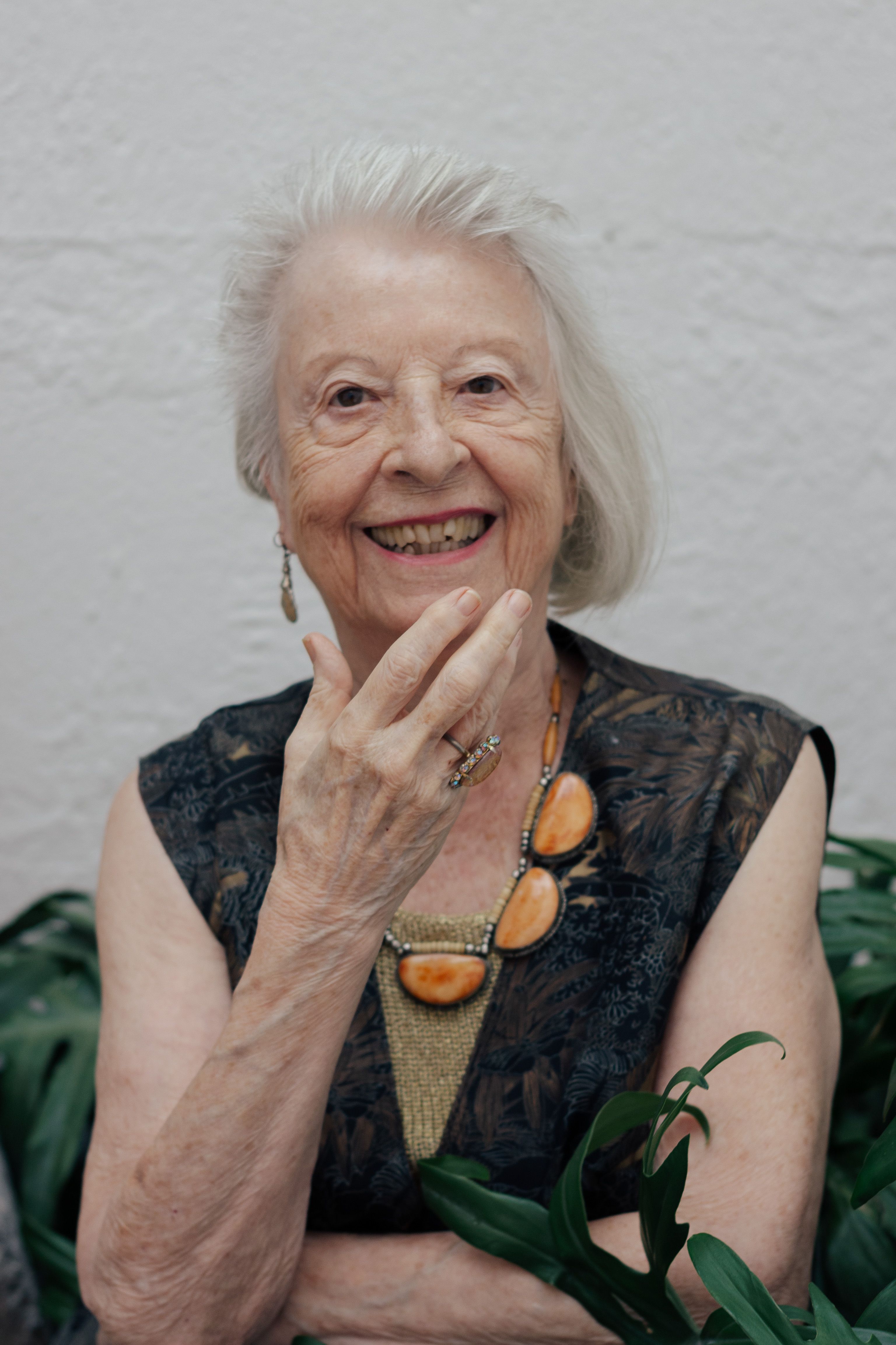 Portrait of a joyful senior woman with elegant jewelry surrounded by greenery indoors.