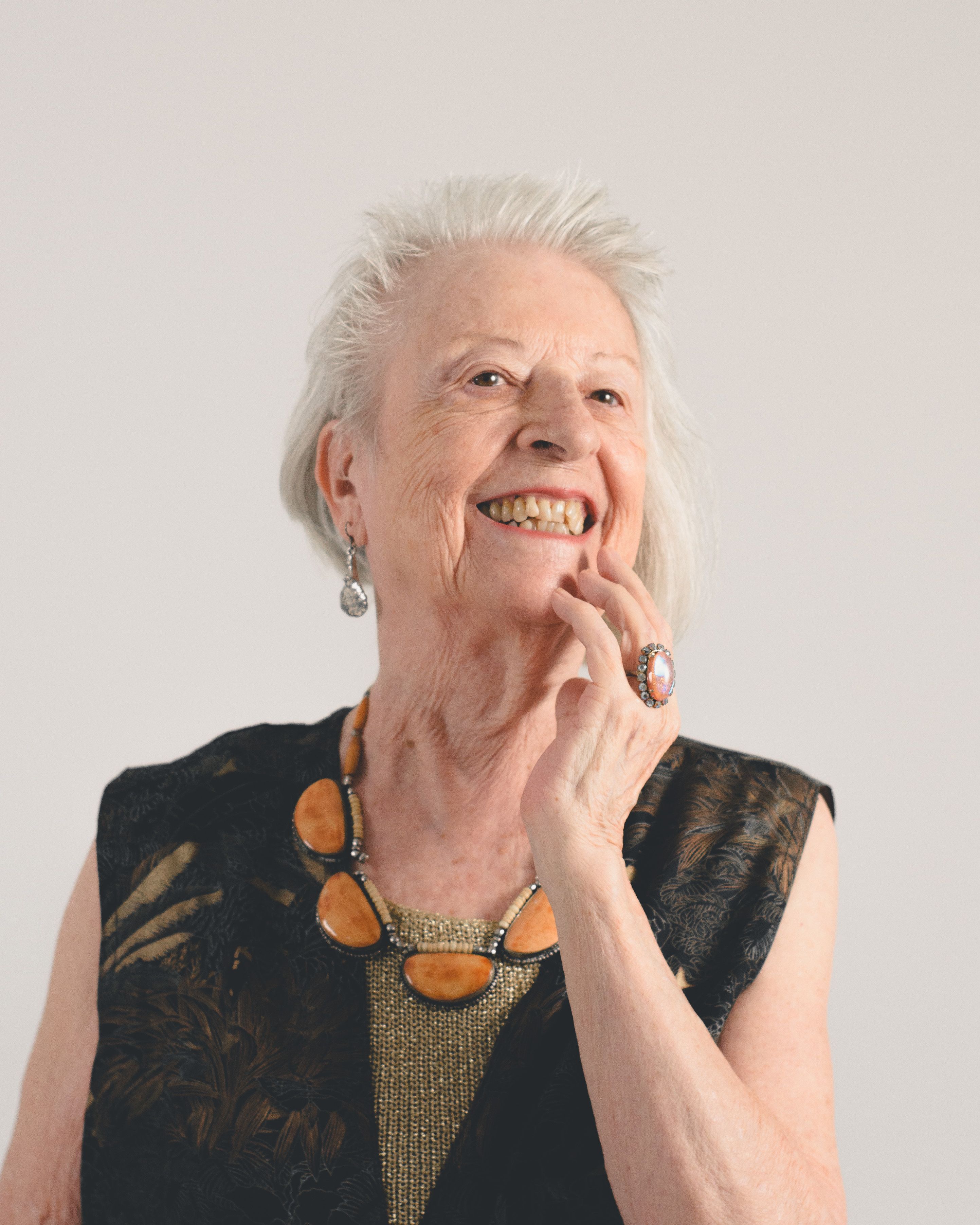 Portrait of a smiling senior woman wearing elegant jewelry and a black dress with a light background.