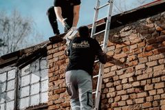 Two construction workers using a ladder to reach a brick building roof under a blue sky.