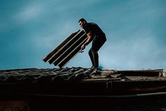 A man on a rooftop repairing with roofing sheets against a deep blue sky.