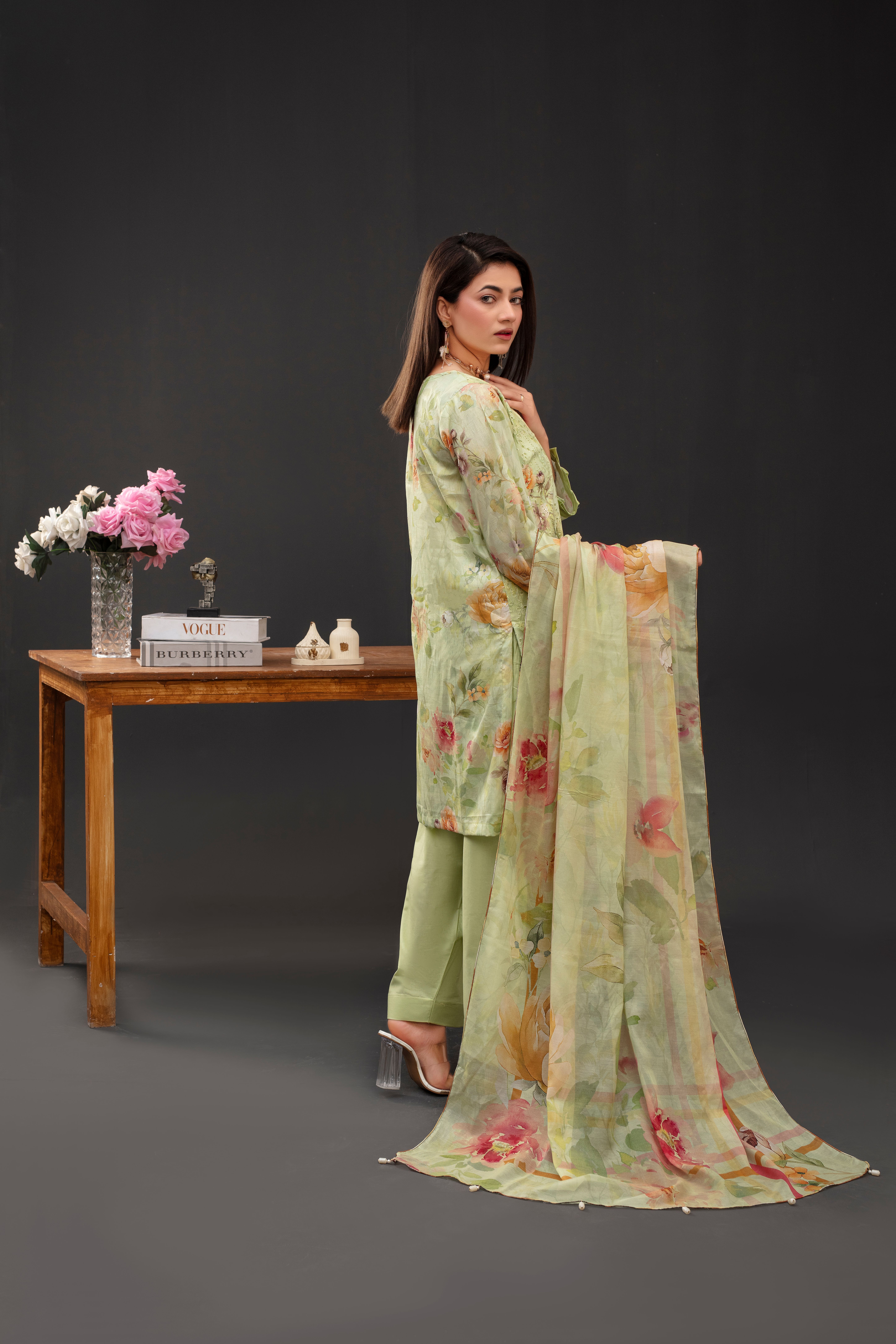 A stylish woman in a floral outfit poses indoors beside a wooden table with flowers.