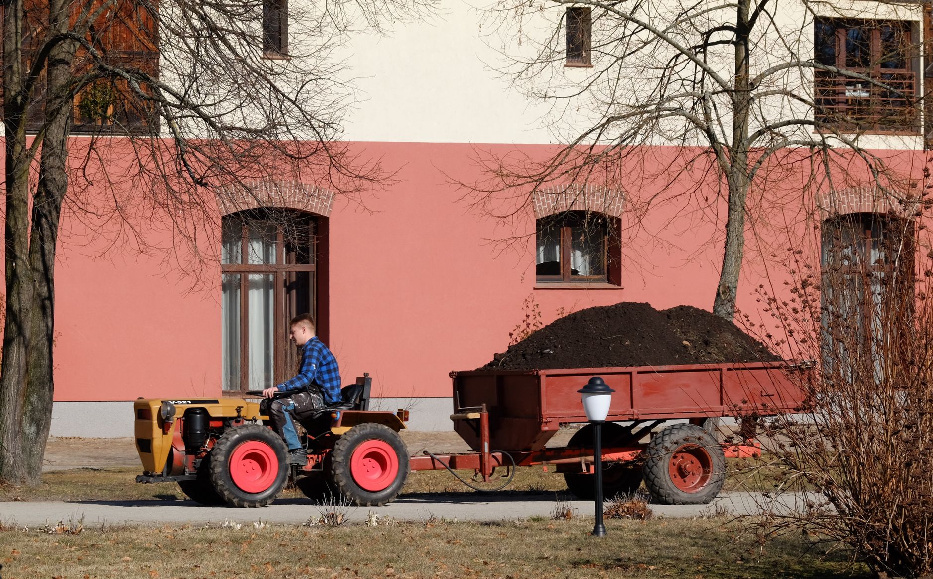 A man in blue plaid shirt drives a tractor pulling a trailer filled with soil near a red building.