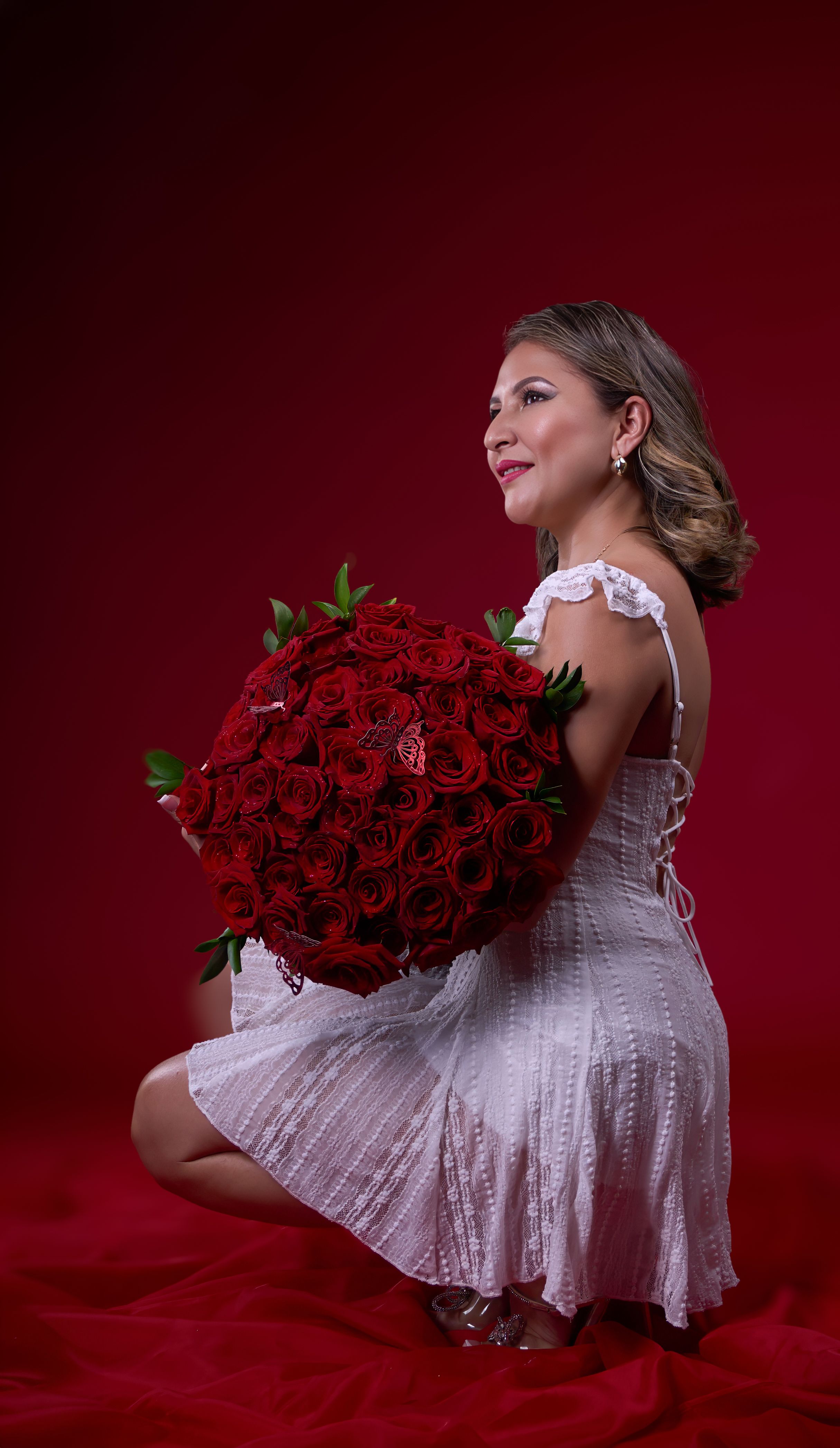A beautiful woman in a white dress holding a large bouquet of red roses, set against a vibrant red backdrop.
