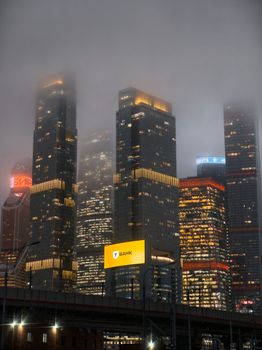 Moody fog enshrouds towering skyscrapers in Moscow's skyline at night, lights warmly glowing.