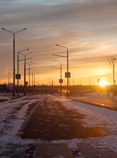 Free A serene sunset casts warm hues over a quiet snow-covered street lined with lampposts. Stock Photo