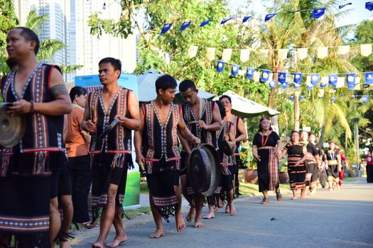 Men and women in traditional attire performing with gongs during a cultural parade outdoors.