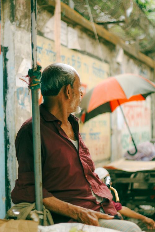 Free Old man sitting by a street pole under an umbrella in the vibrant streets of Old Dhaka. Stock Photo