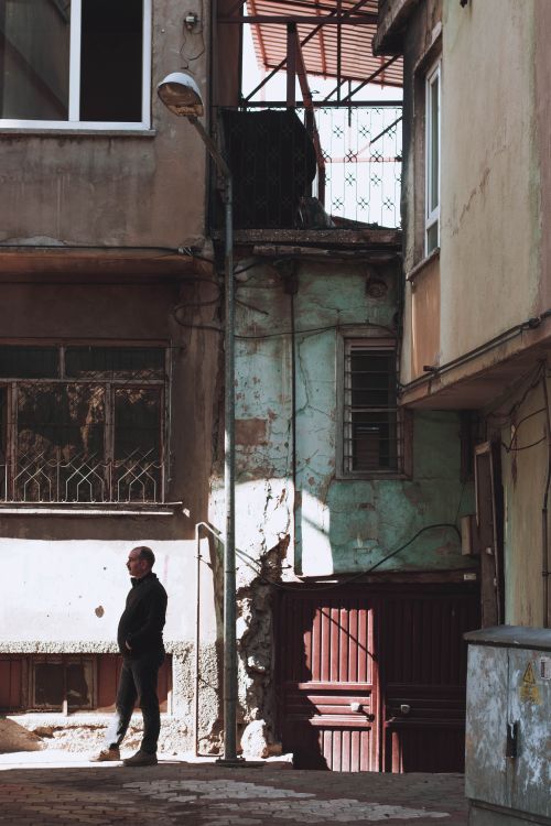 Free A man stands in a shadowy urban alley surrounded by aged, rustic buildings. Stock Photo