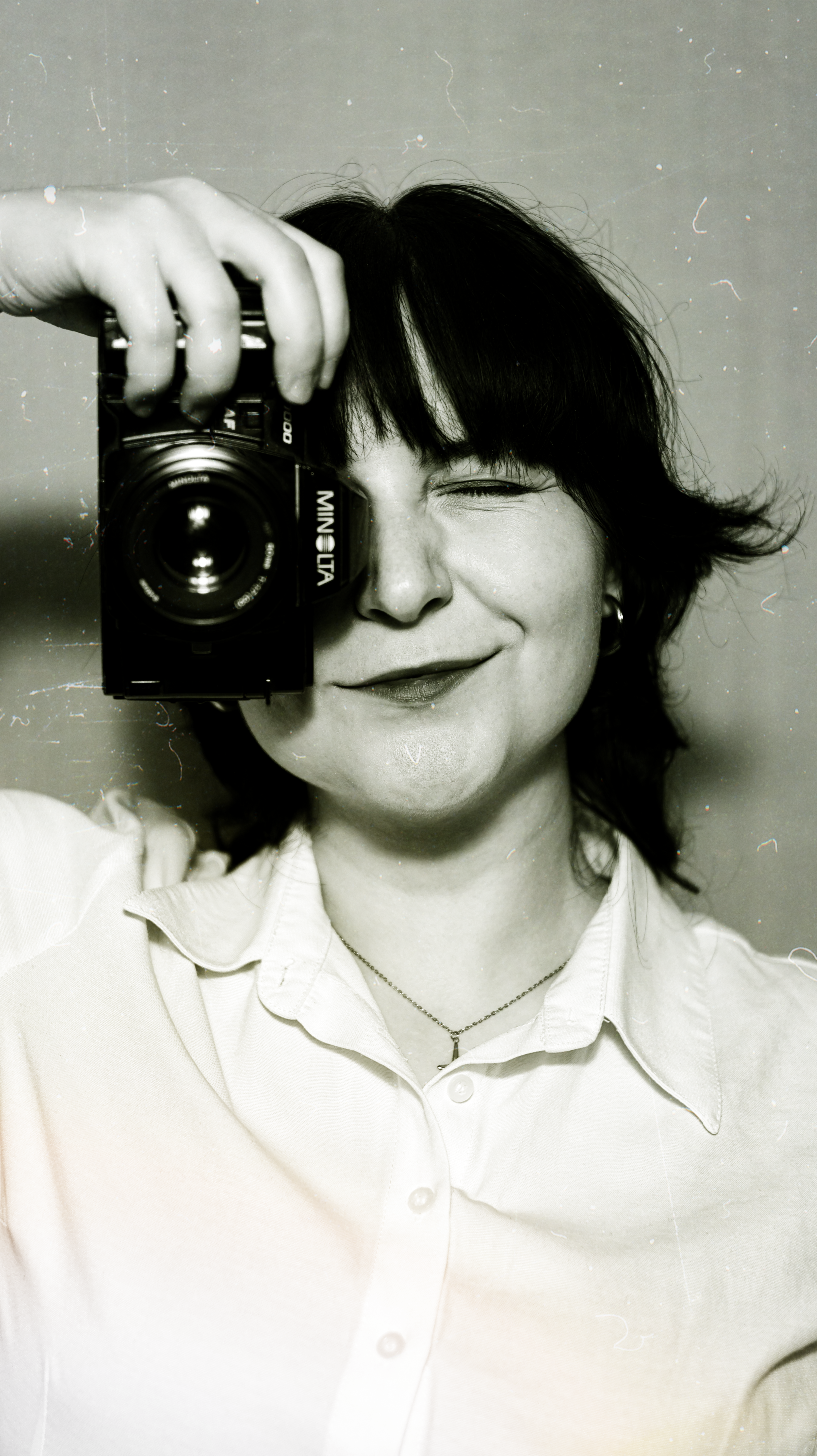 A black and white portrait of a smiling woman holding a camera, exuding a vintage charm.