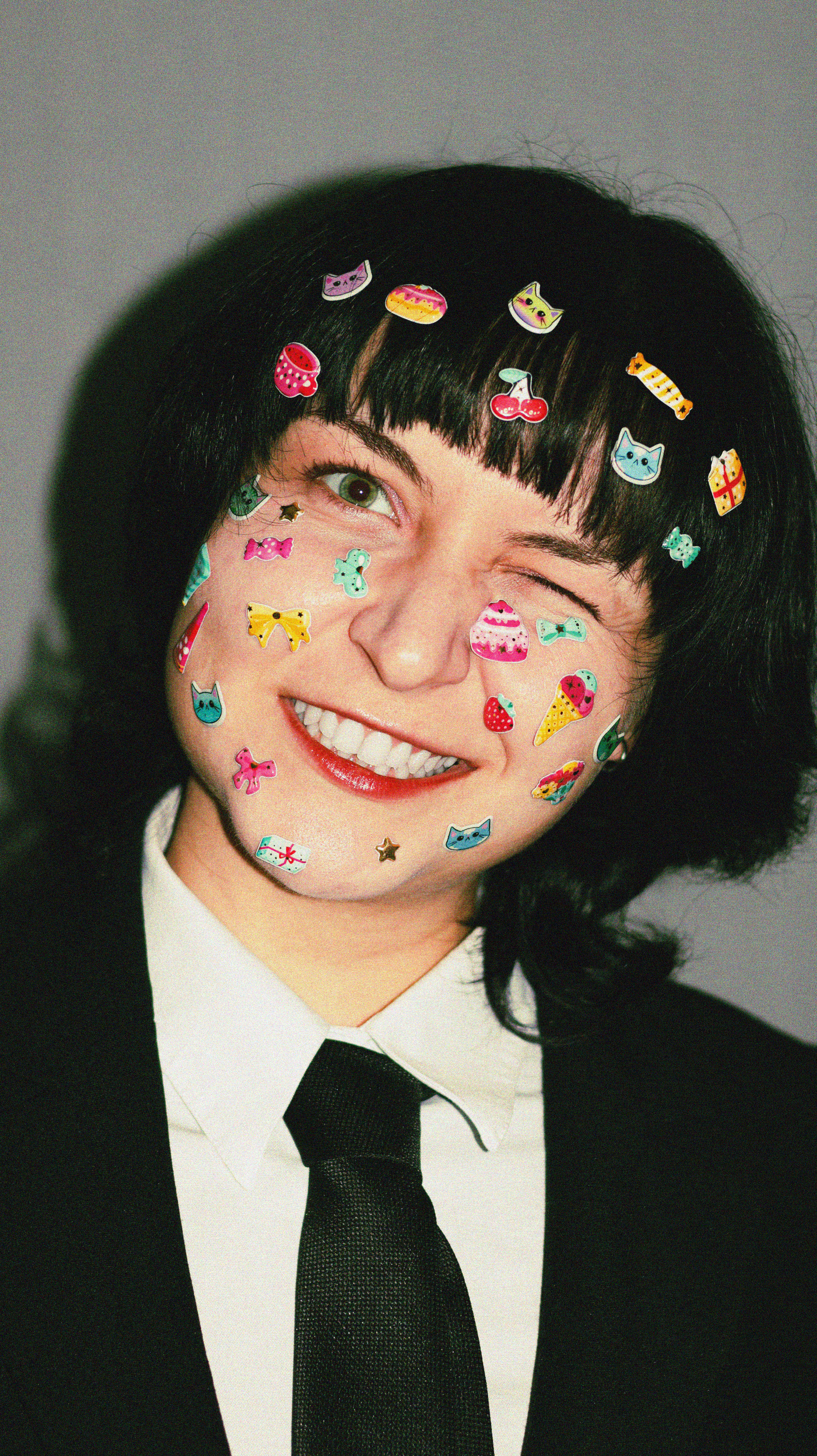 Smiling adult with black hair covered in colorful stickers. Playful and quirky expression.
