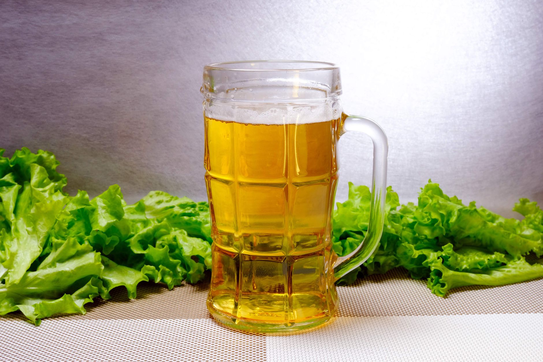 A glass mug of golden beer on a checkered table with lettuce around. Vibrant and refreshing.
