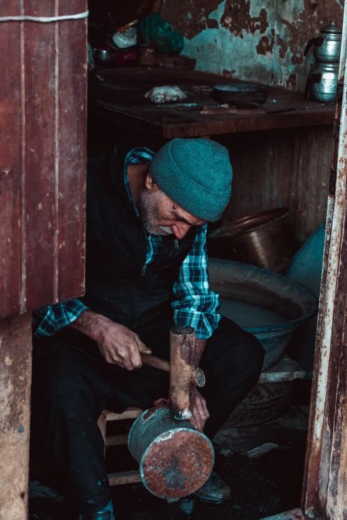 Free Mature poor male in worn out clothes sitting in weathered messy outbuilding and fixing metal pan with self made wooden hammer Stock Photo