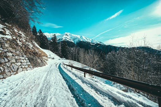 A tranquil winter scene featuring a snow-covered road winding through a scenic mountain landscape under a clear blue sky.