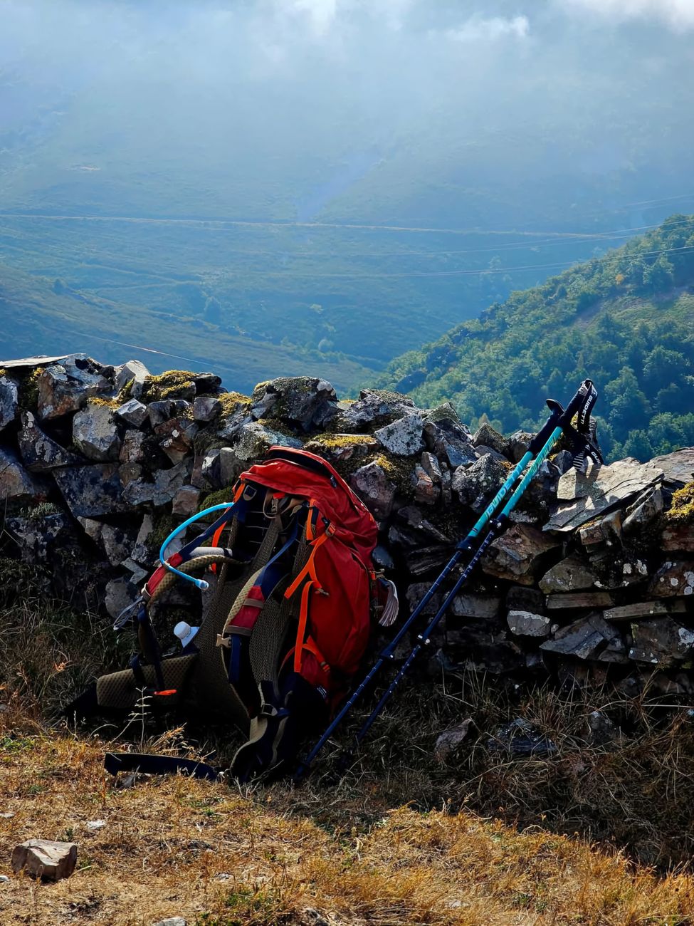 Un sac à dos rouge avec des bâtons de randonnée adossé à un mur de pierre surplombant un paysage montagneux luxuriant.