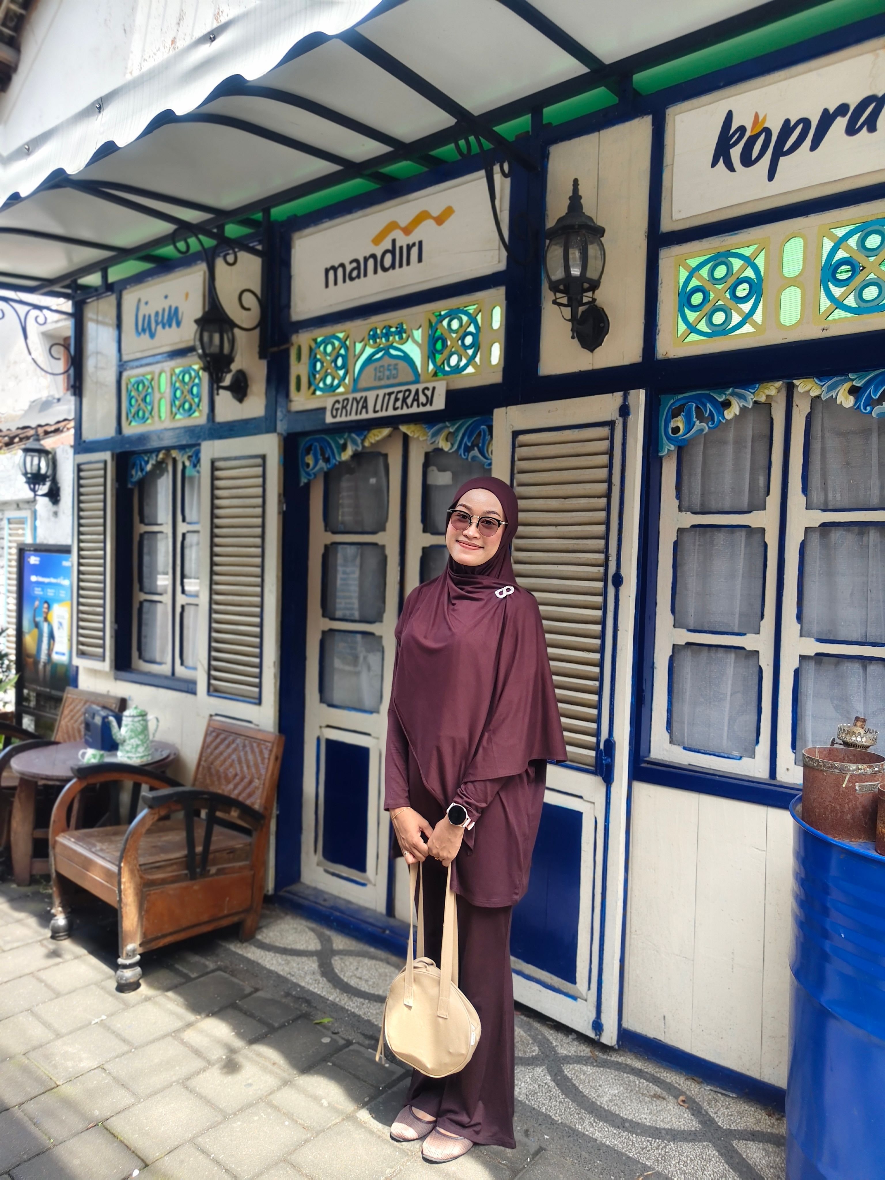 A woman in a hijab poses outside Griya Literasi, Java. Bright daylight enhances the cultural setting.