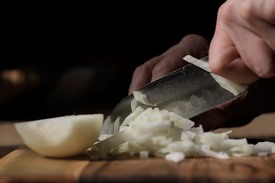 A person skillfully slices onions with a knife on a wooden board in a kitchen setting.