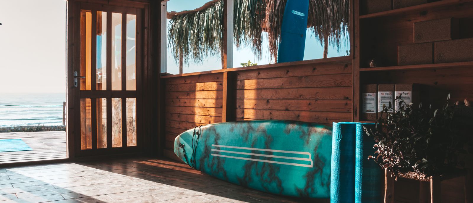 Wooden surf shack interior with surfboard and yoga mat overlooking the ocean at sunrise in Taghazout, Morocco.
