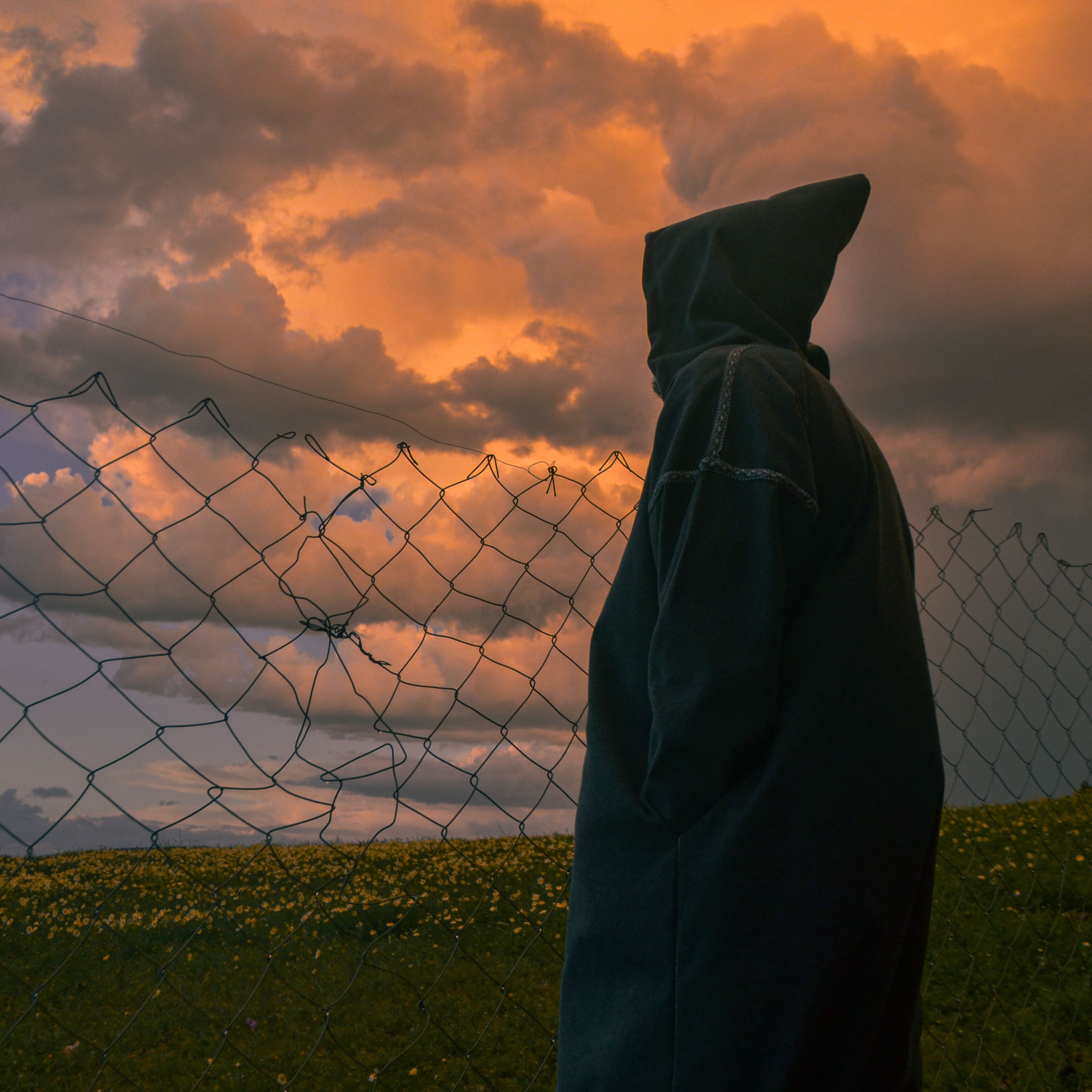 A dramatic silhouette of a hooded figure standing by a fence under a vibrant sunset sky.