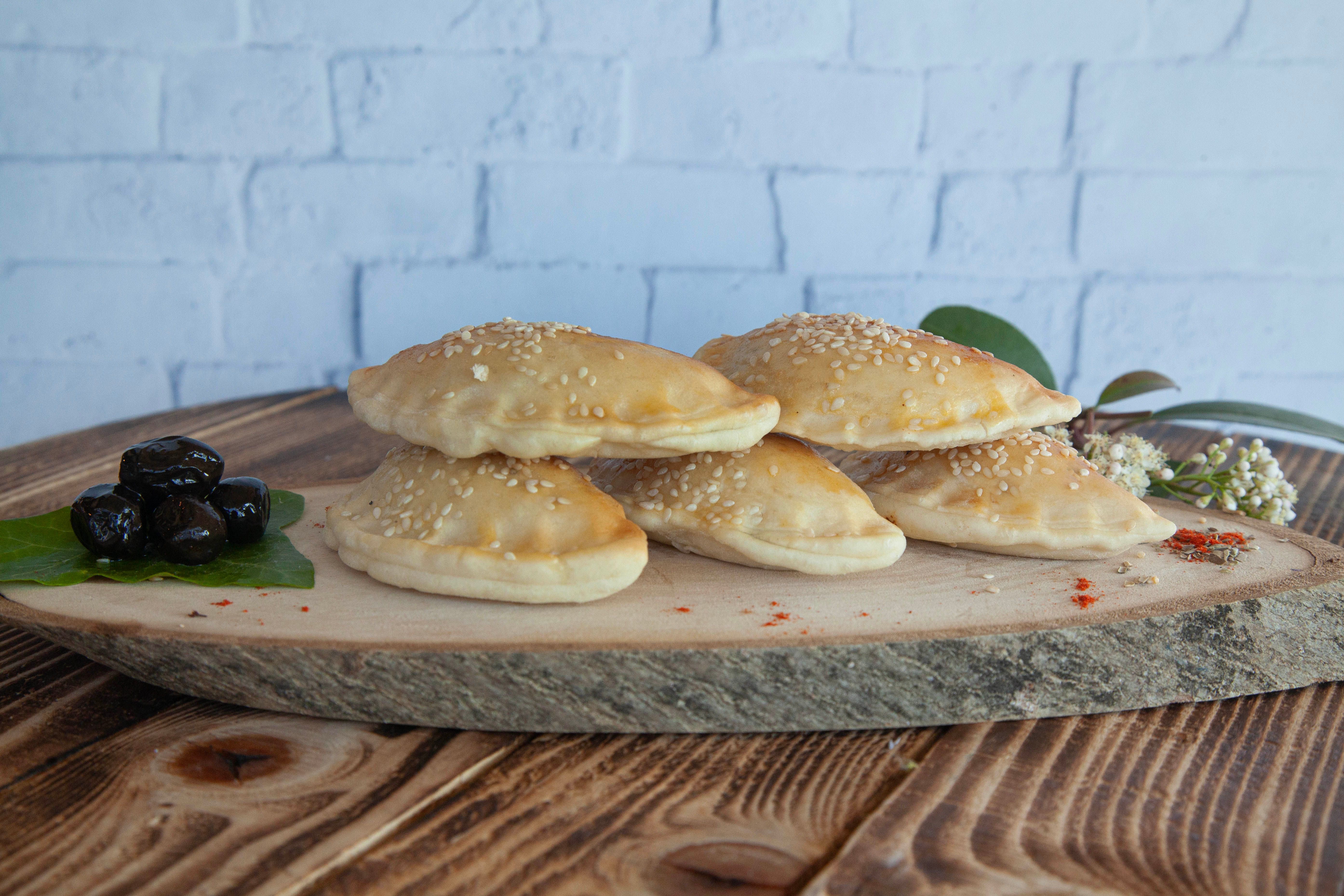A stack of sesame-topped pastries displayed on a rustic wooden board with olives and seasoning.