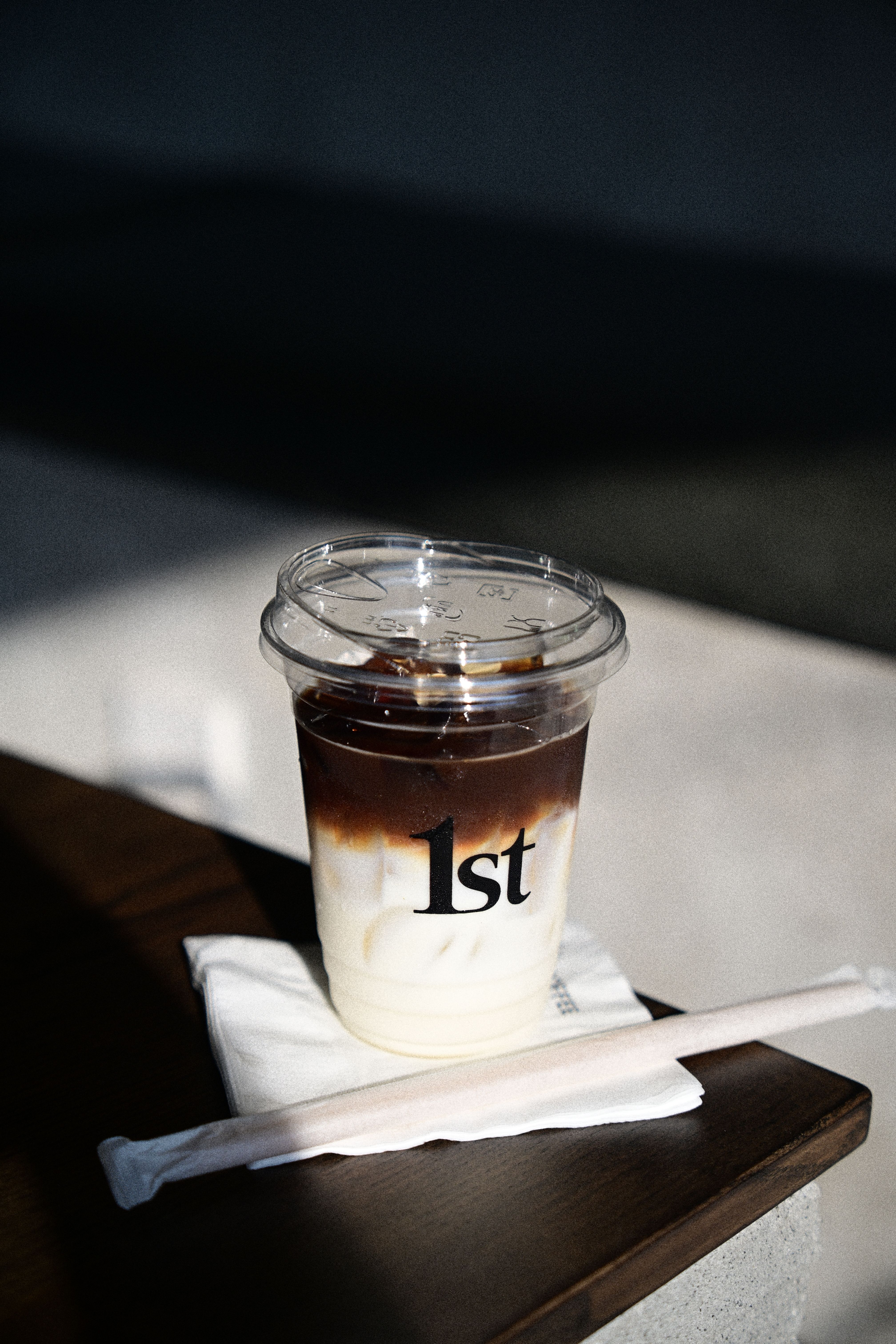 Close-up of iced coffee in a plastic cup with '1st' logo on a table in sunlight.