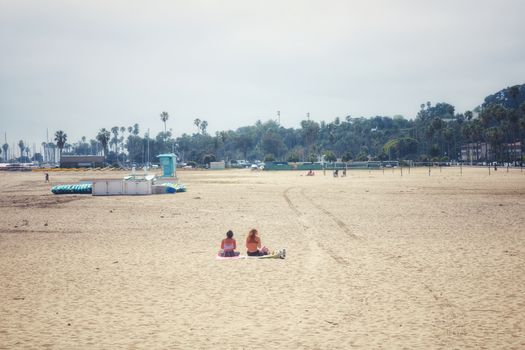 Two people relax on Santa Barbara beach, CA, with palm trees and lifeguard tower.
