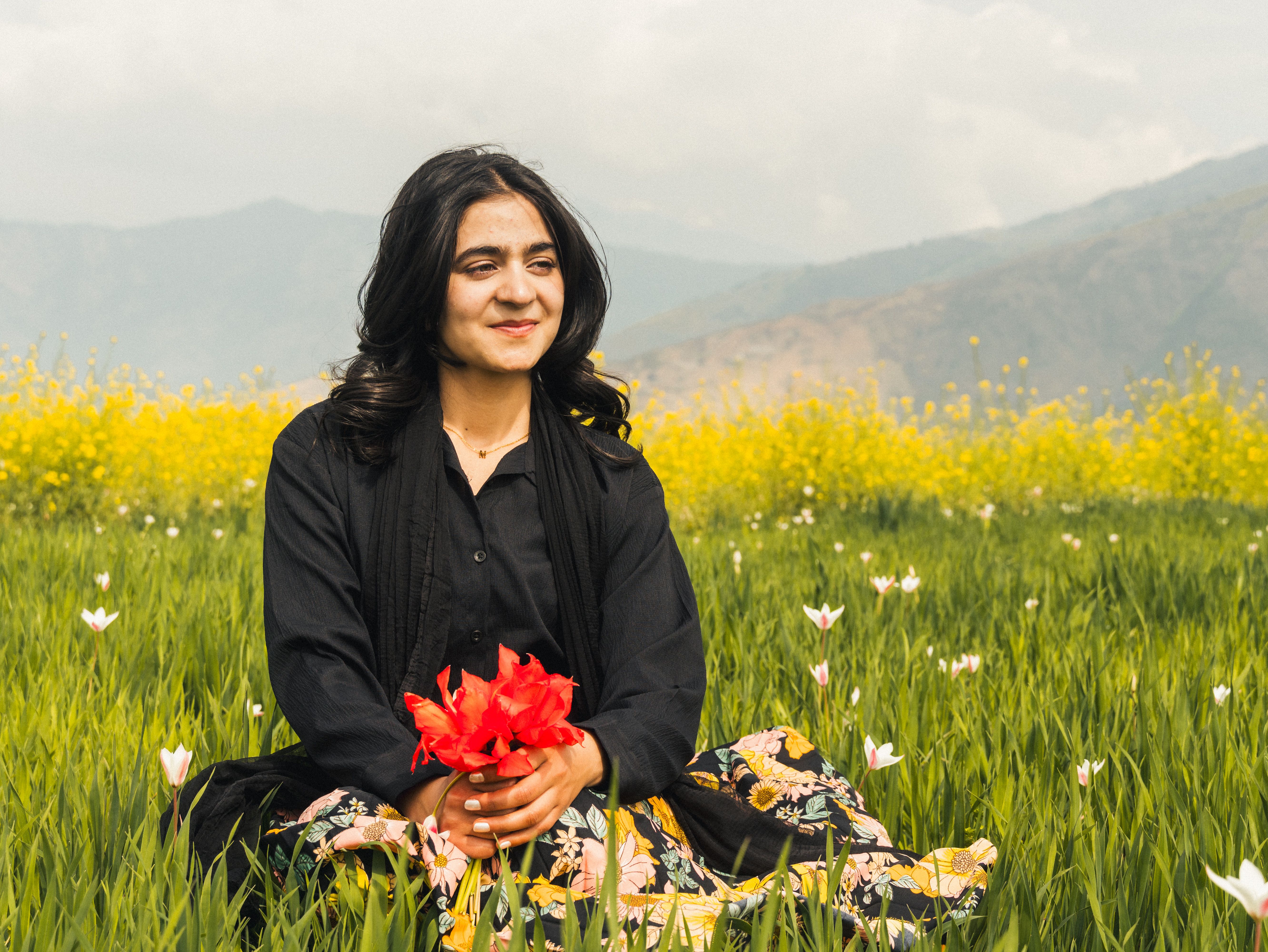 Smiling woman with flowers sitting in a vibrant meadow with mountains in the background.