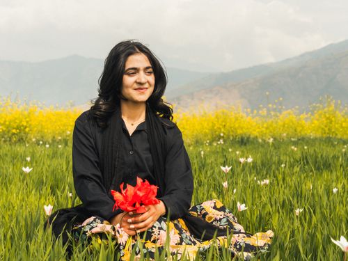 Free Smiling woman with flowers sitting in a vibrant meadow with mountains in the background. Stock Photo