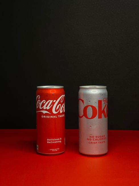 A silver Diet Coke can and a black Coke Zero Sugar can standing next to each other on a countertop, both with condensation beading on the metal