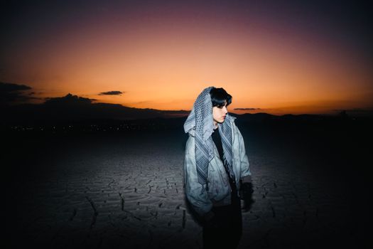 A young man in a hoodie stands in a desert under a vibrant sunset sky, creating a moody scene.