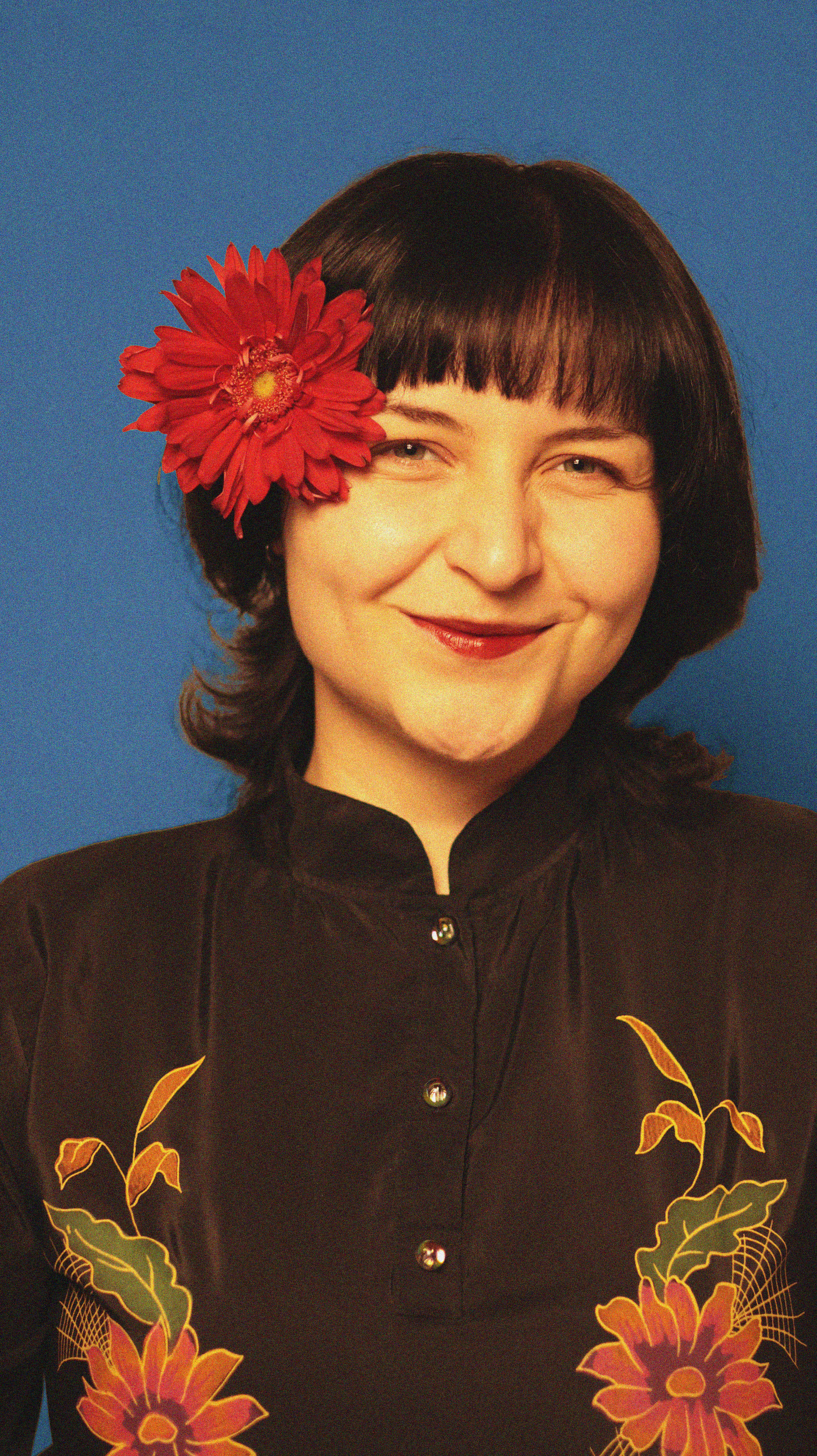 Portrait of a smiling woman with a red flower in her hair against a blue background.
