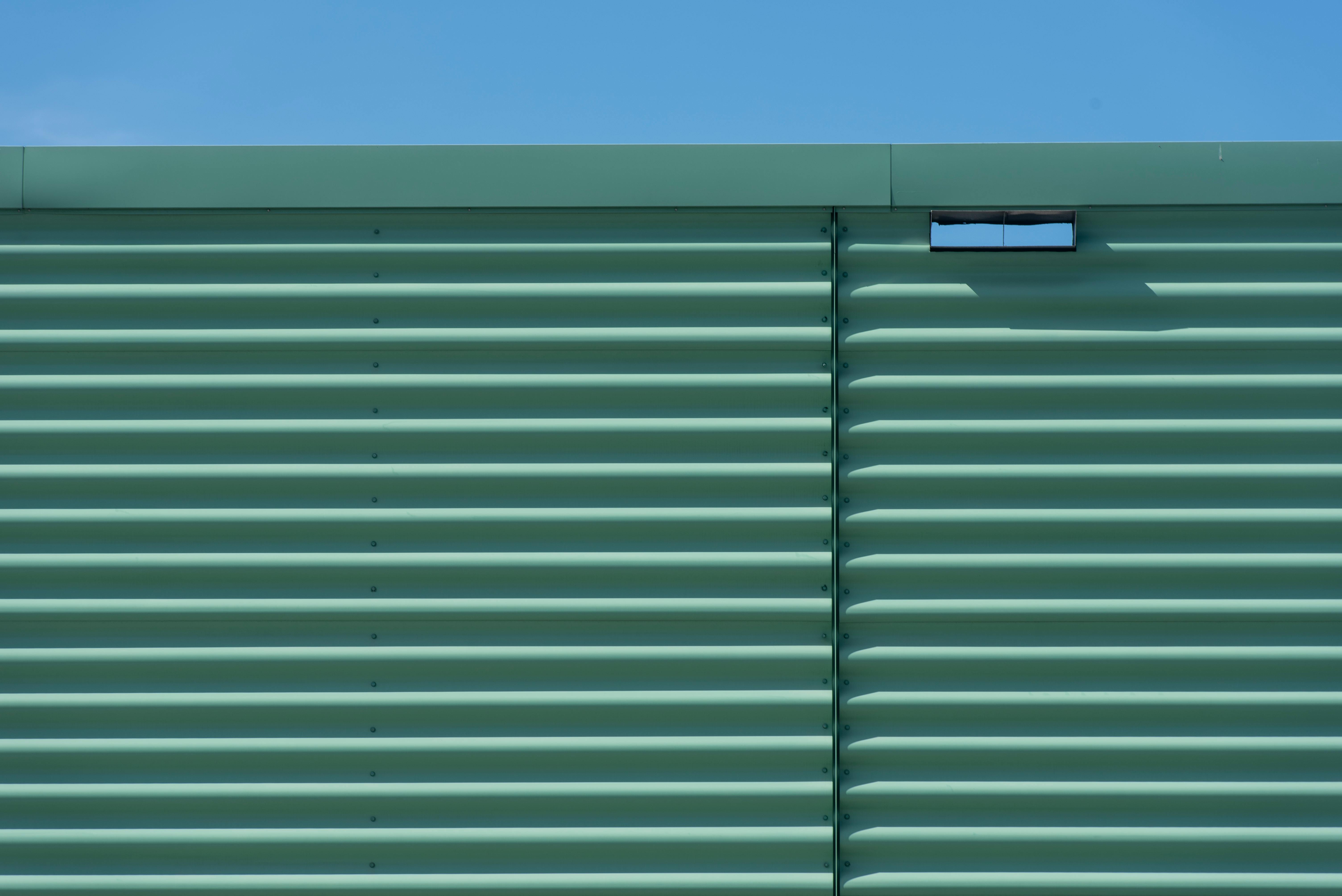 Vue minimaliste d'un mur métallique vert à panneaux horizontaux se détachant sur un ciel bleu clair, prise en extérieur.