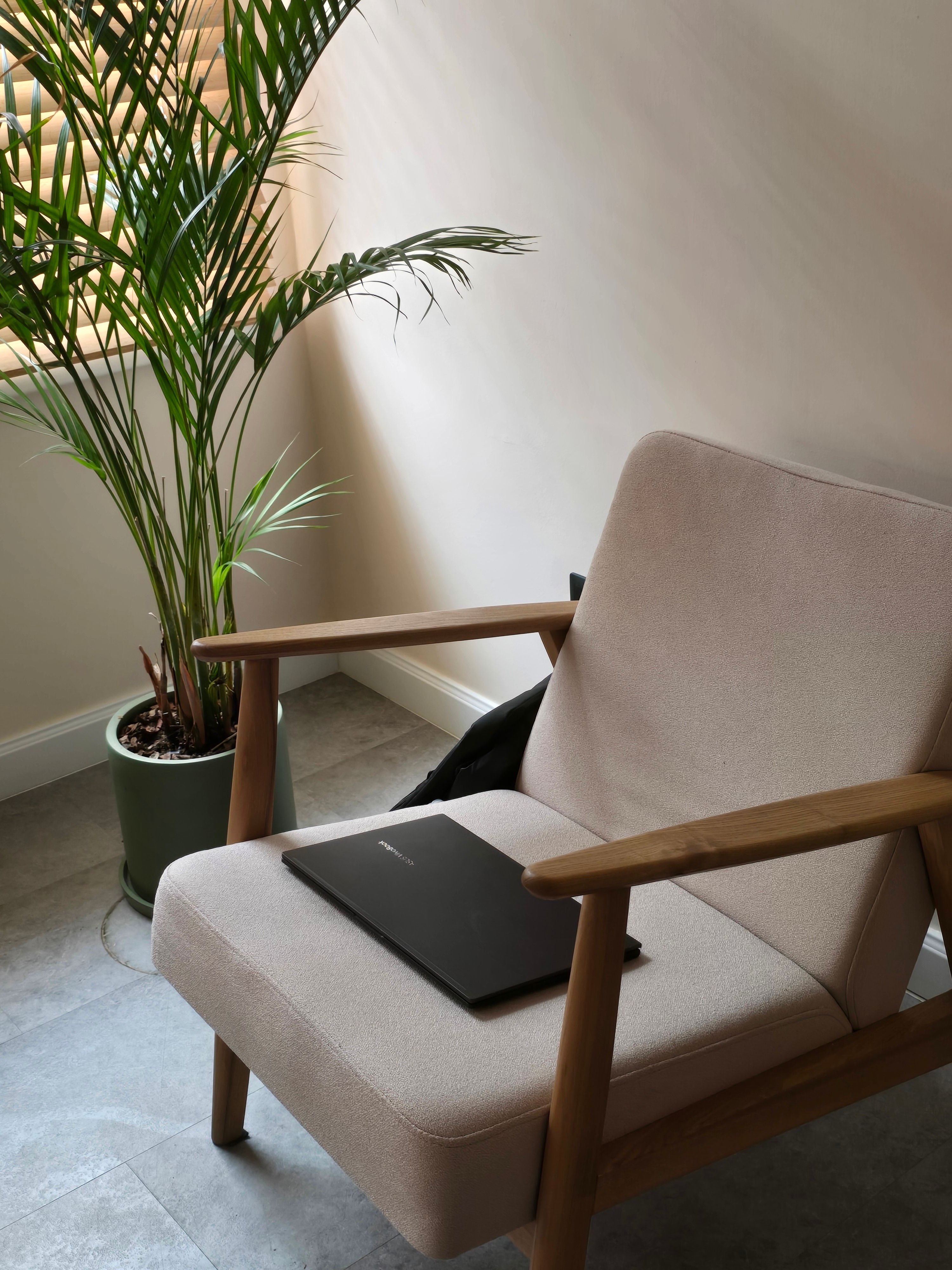 Comfortable armchair with laptop placed on it beside a potted plant in a sunlit modern room.