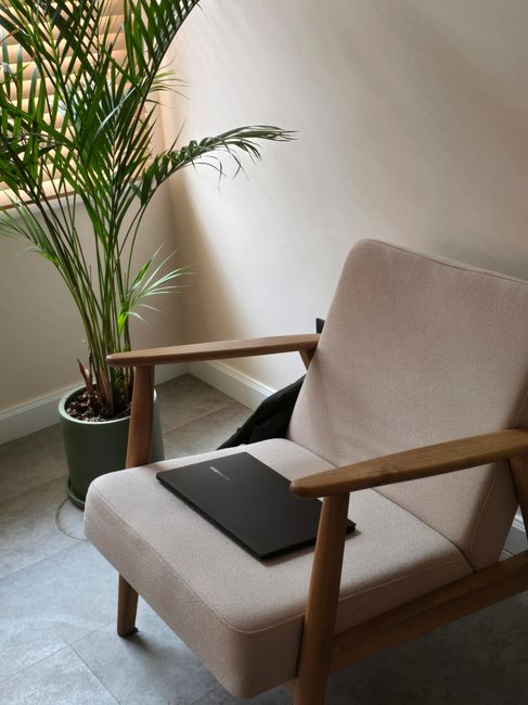 Comfortable armchair with laptop placed on it beside a potted plant in a sunlit modern room.