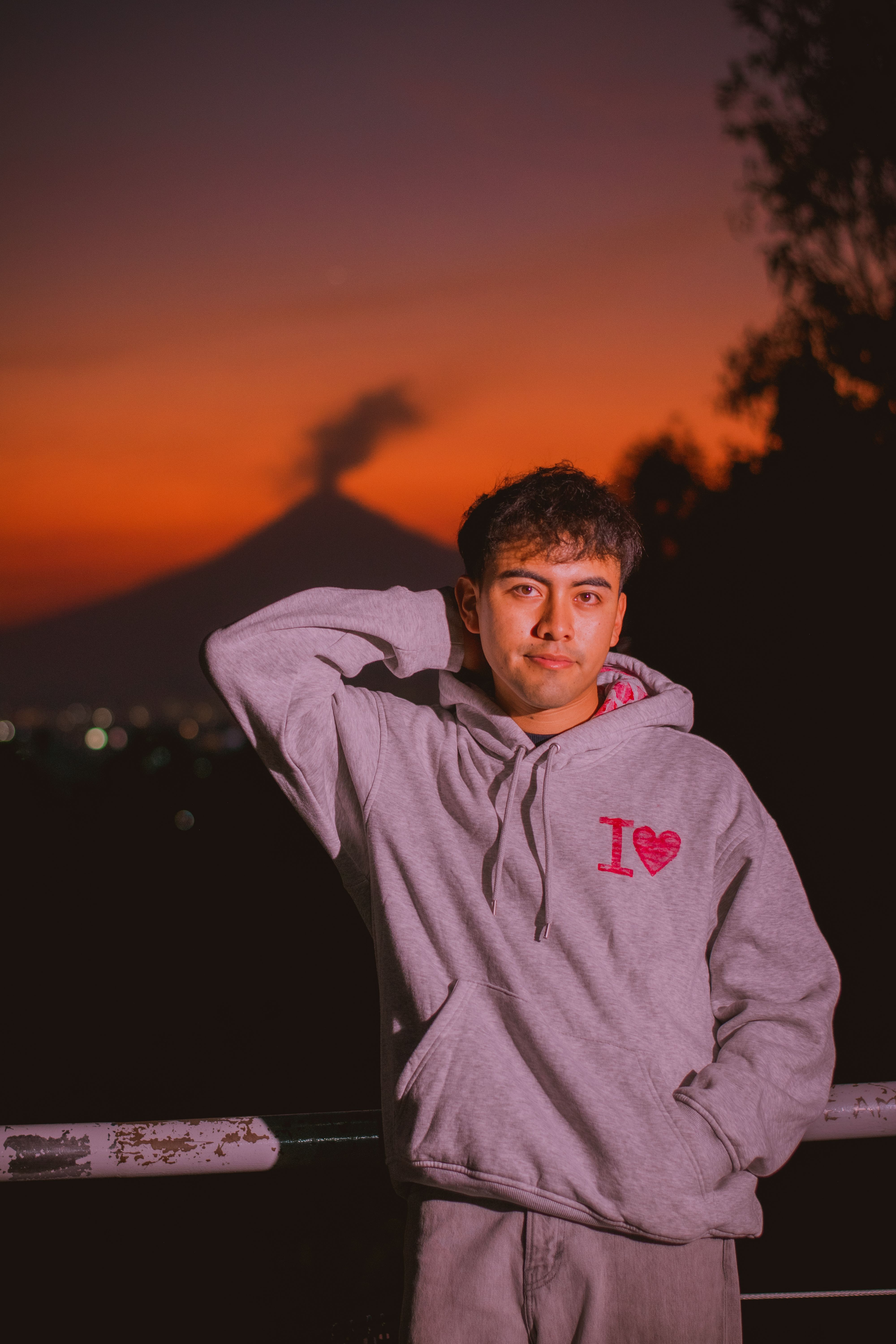 Young man in a hoodie poses at sunset with a volcano in the background, creating a dramatic silhouette.