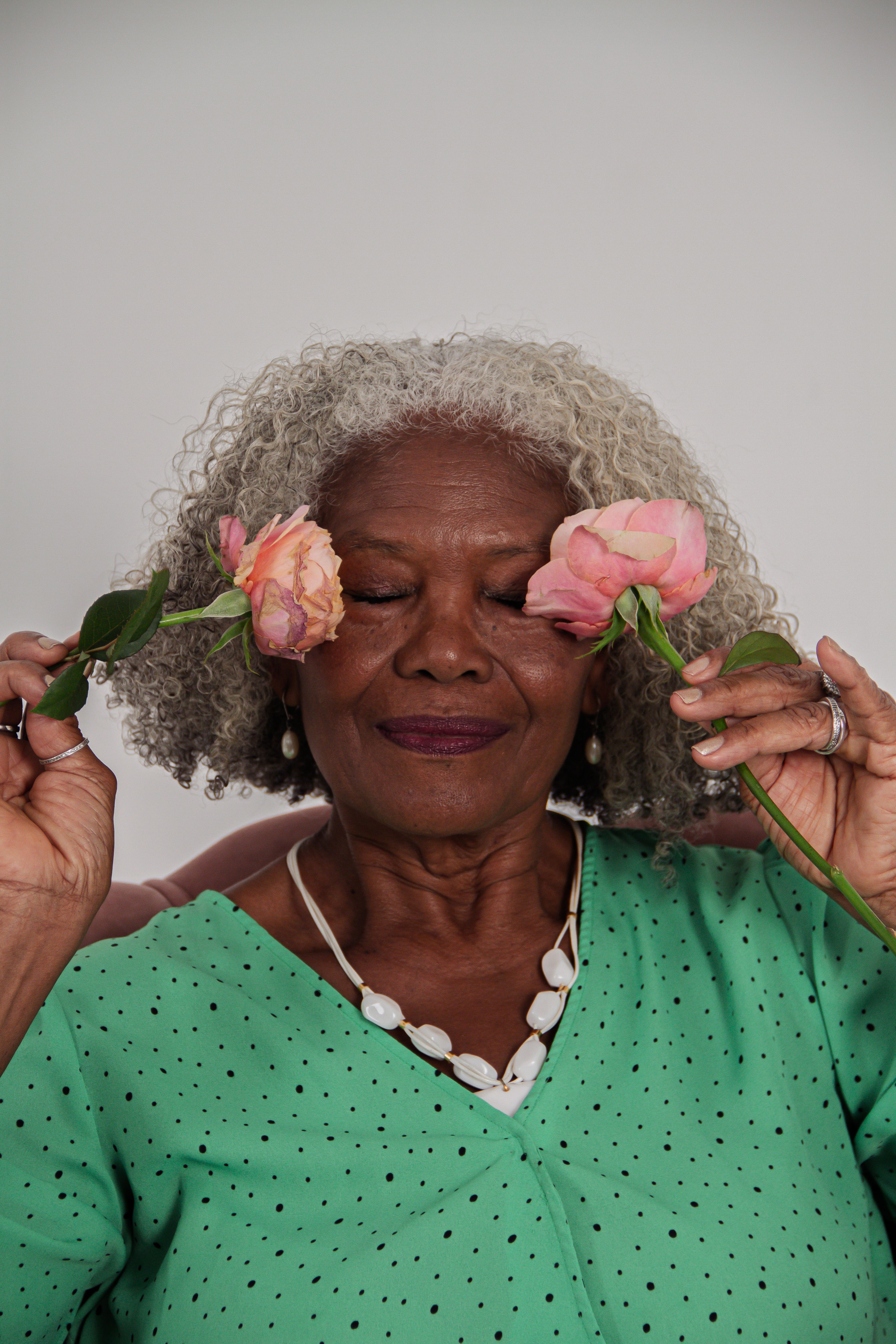 Elegant senior woman with gray hair playfully holding pink roses to her eyes.