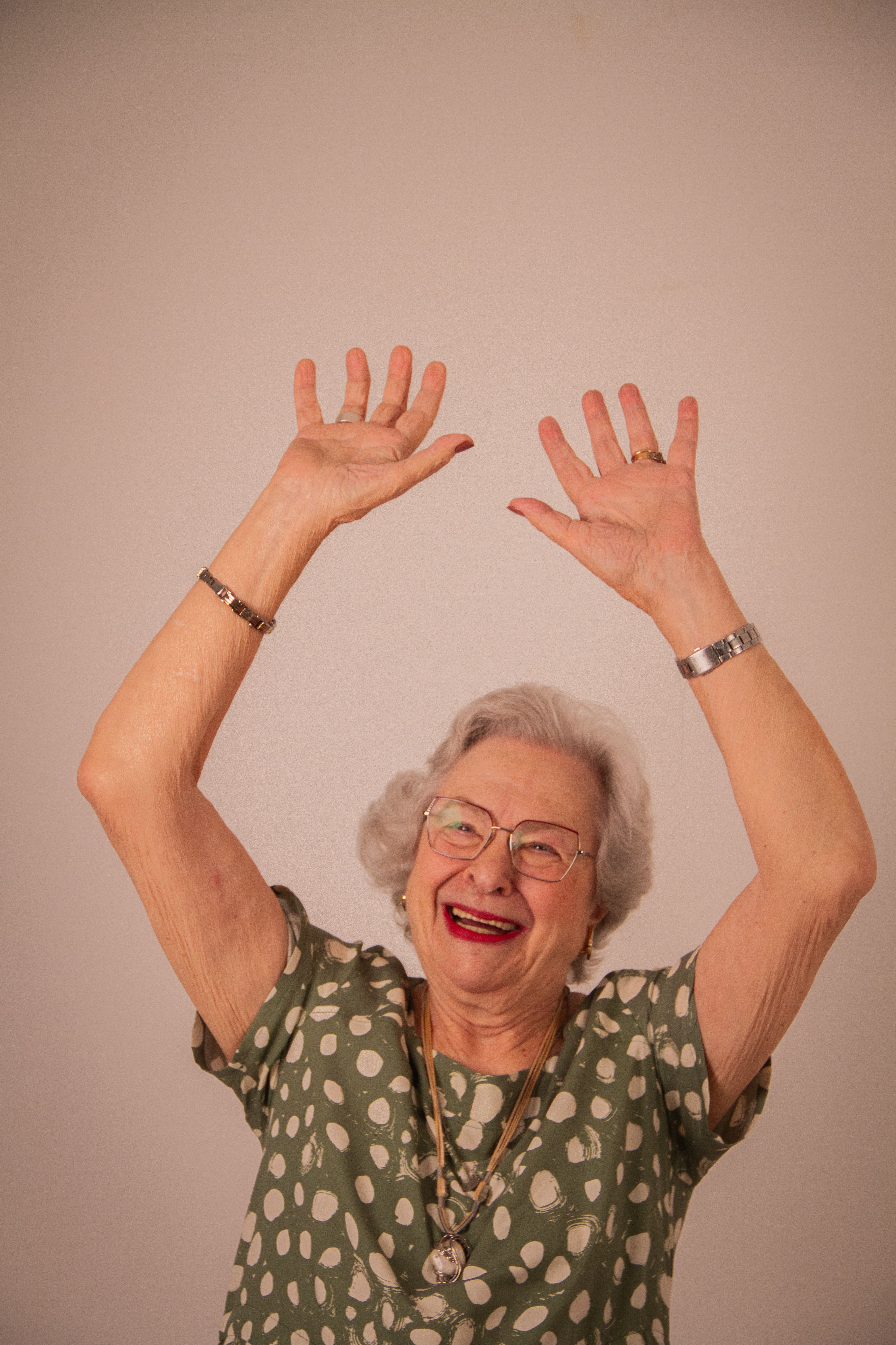 Elderly woman in a green dress with polka dots raising hands joyfully indoors.