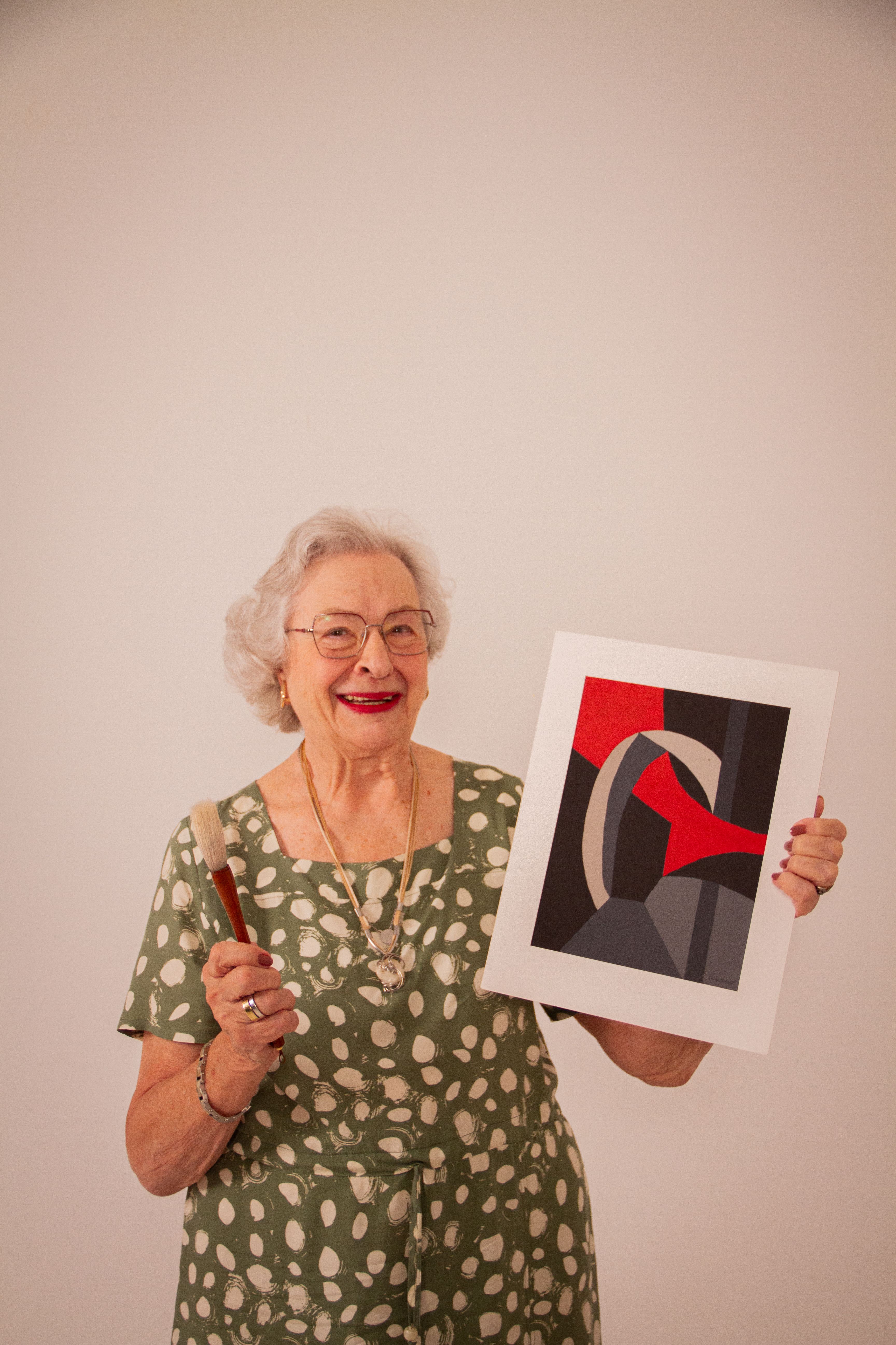 Senior woman with gray hair holding an abstract painting and brush, smiling in São Paulo.