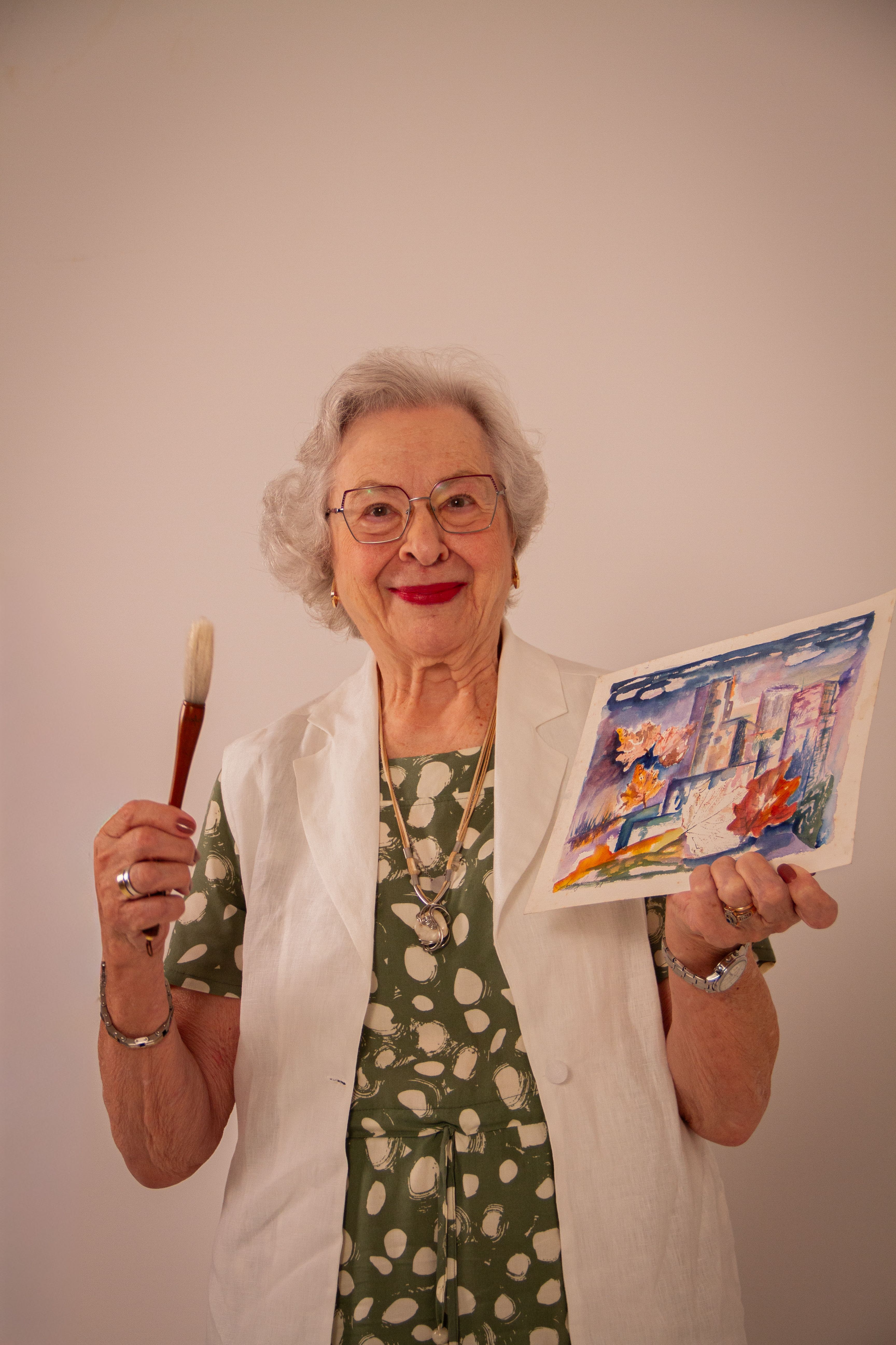 Senior woman artist joyfully holding watercolor painting and brush with a smile.