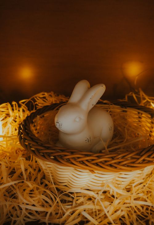 Free Charming ceramic bunny in a wicker basket surrounded by warm lights and straw, perfect for Easter decor. Stock Photo