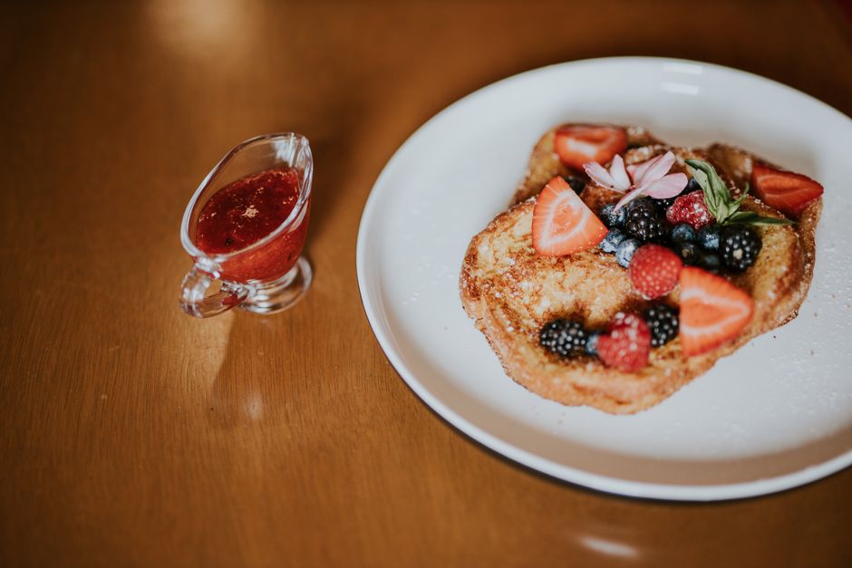Tasty French toast topped with fresh berries and served with a side of berry syrup on a wooden table.