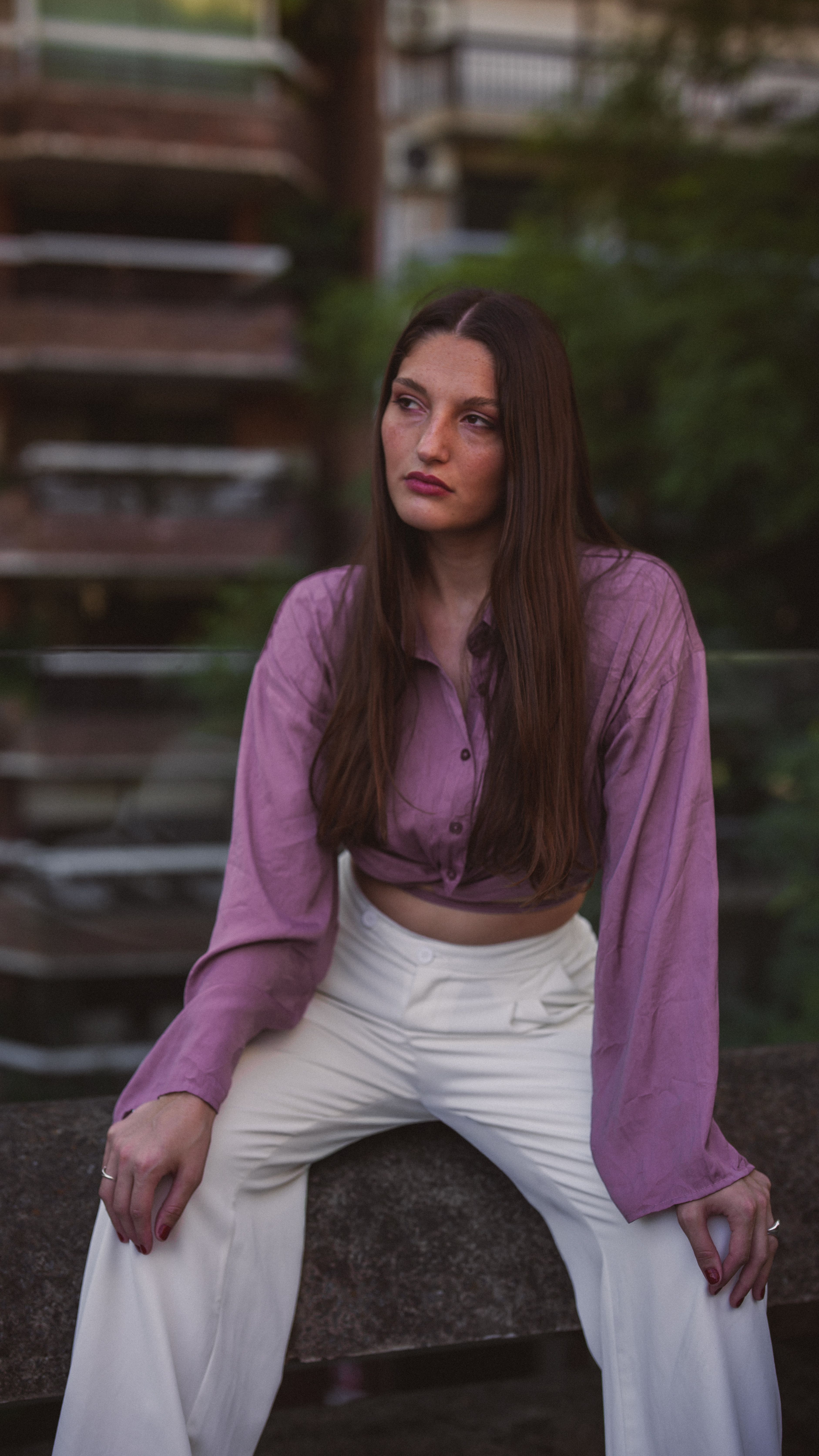 Young woman with long hair wearing a chic outfit sitting outdoors with a contemplative expression.