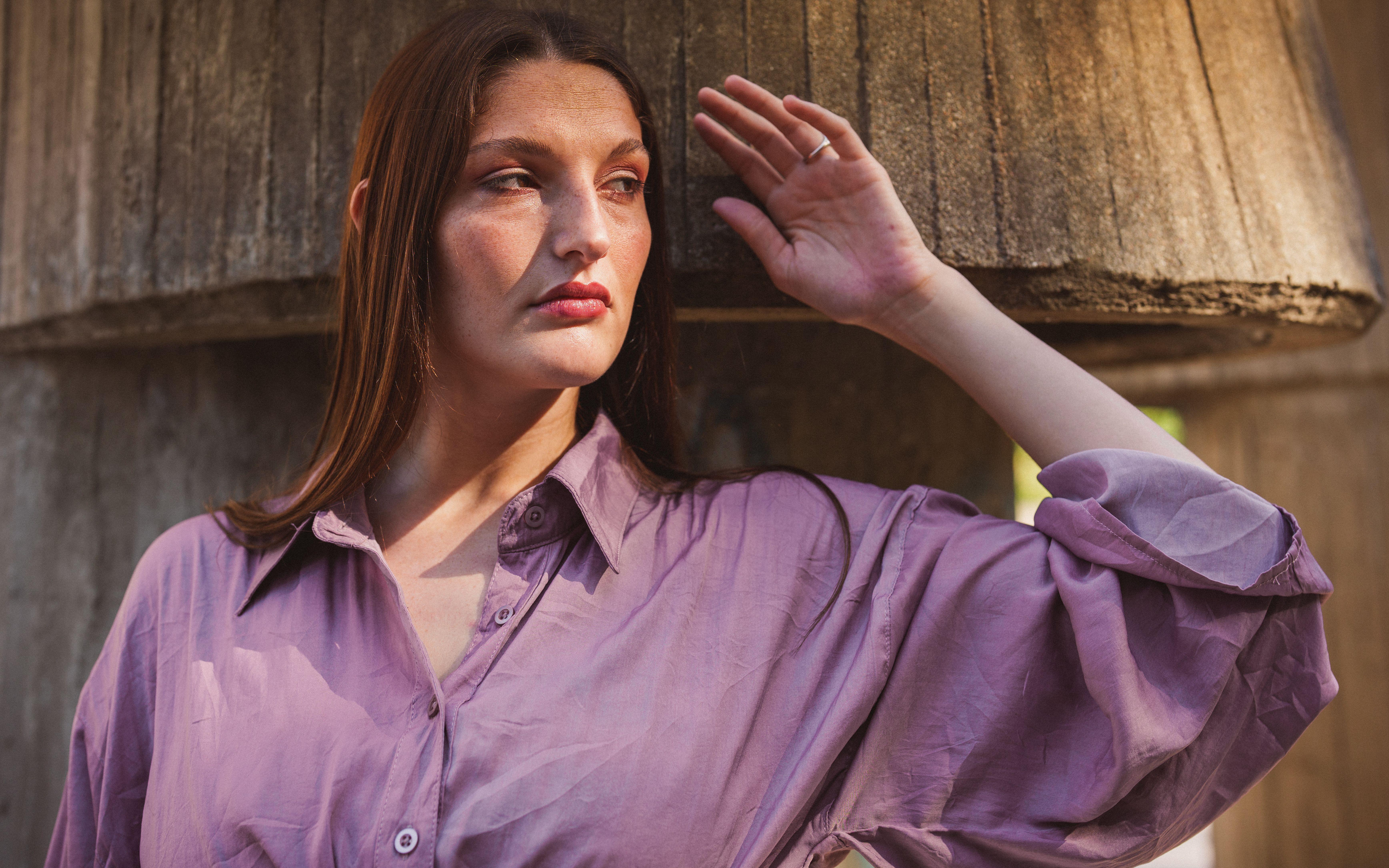 A thoughtful woman in a purple shirt poses against a rustic background, highlighting natural light and textures.