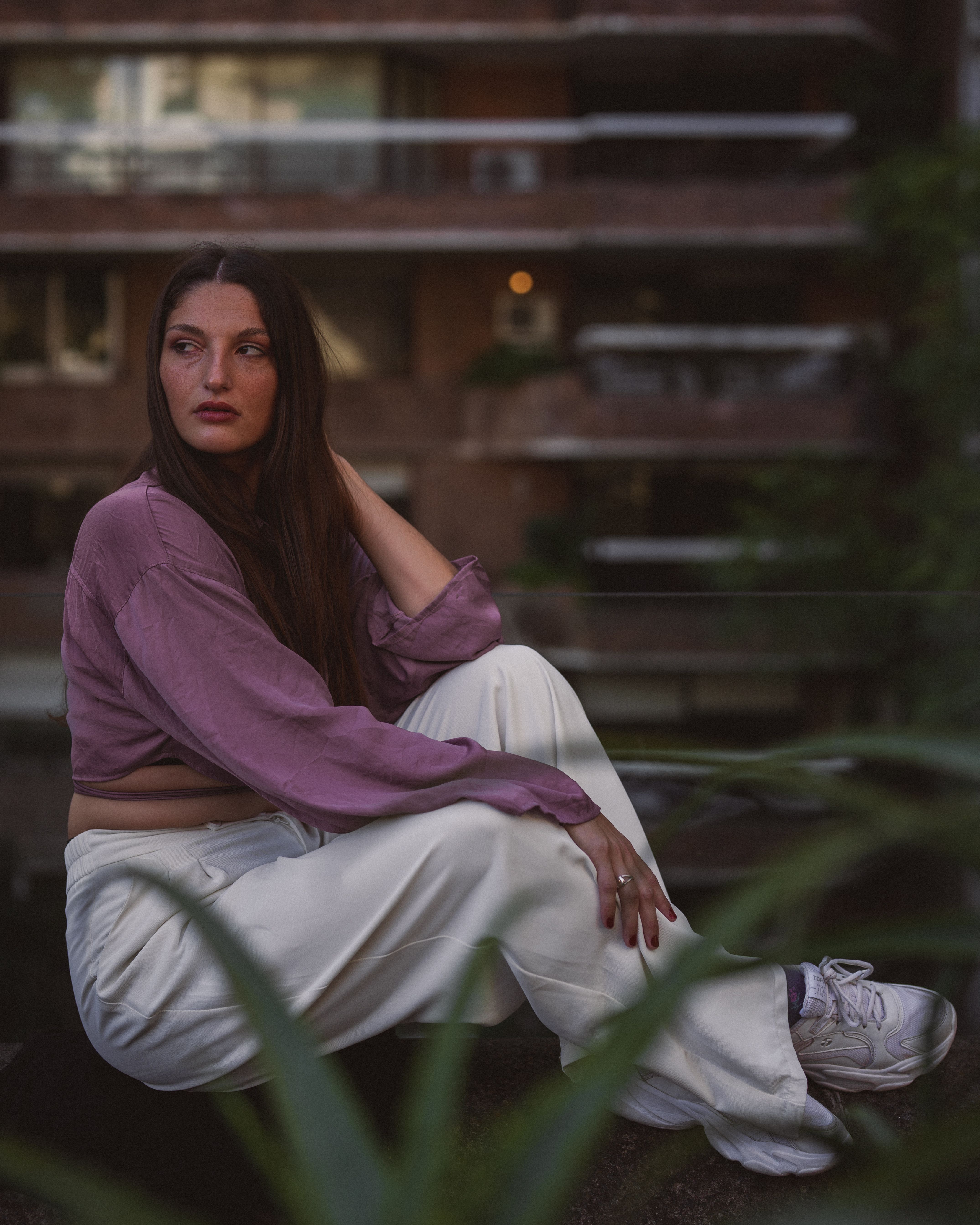 Young woman in casual wear sitting outdoors in front of a city apartment building.