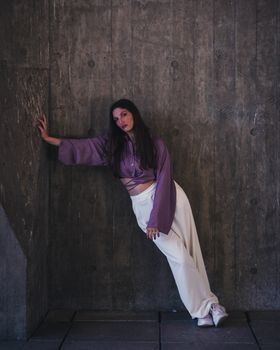 Fashion model in purple top and white pants leans dramatically on a concrete wall.