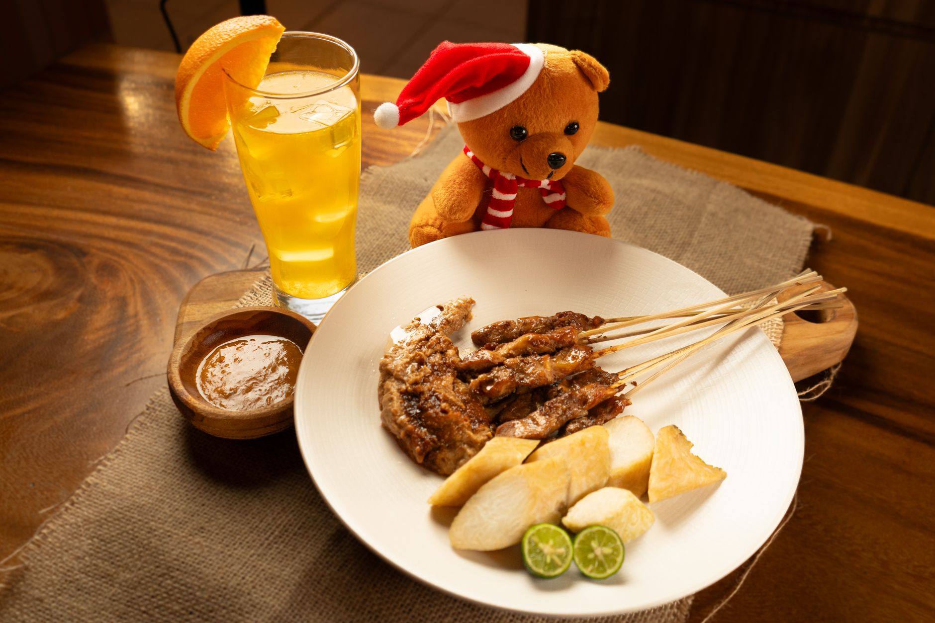A festive plate of chicken satay with a teddy bear, served with citrus drink for Christmas celebration.