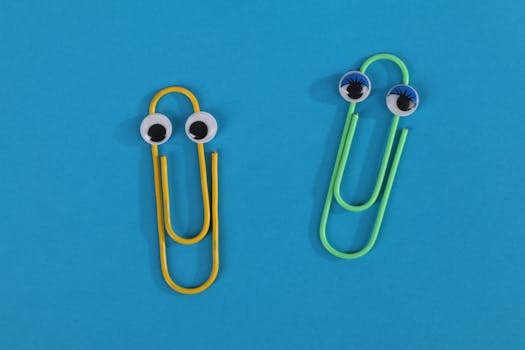 Whimsical image of yellow and green paperclips with googly eyes on a blue surface, creating a playful office theme.
