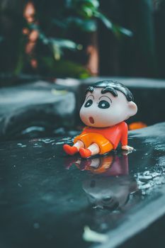 Close-up of a cartoon figure sitting on a wet surface with reflection, outdoors.