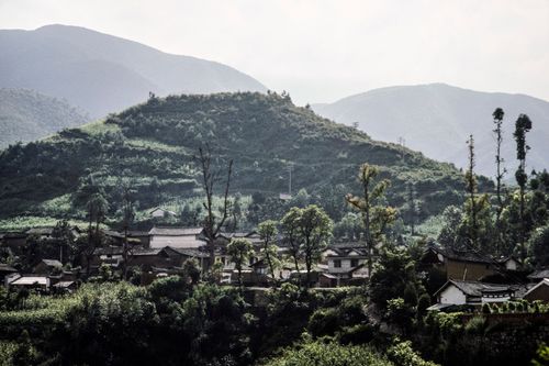 Free Charming village nestled among lush green hills with mountain backdrop and clear sky. Stock Photo